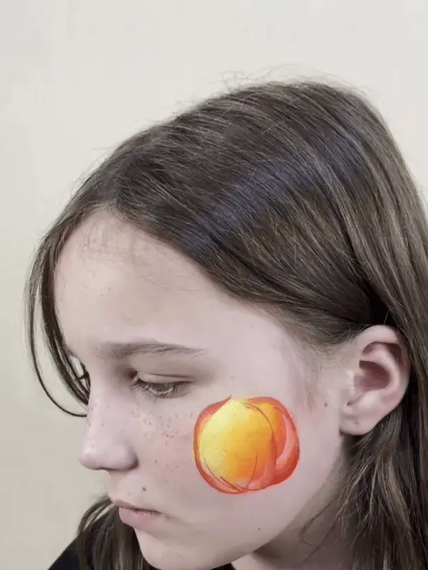 girl has an unfinished painting of a organge pumpkin on her left cheek
